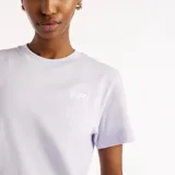 Remera New Balance modelo Sport Essentials para mujer, de color lila con cuello redondo, mangas cortas y calce clásico. Presenta un pequeño logo de la marca bordado en el pecho.