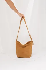 Cartera tipo morral de gamuza color tostado, con correa larga ajustable con hebilla metálica y diseño de base cuadrada.