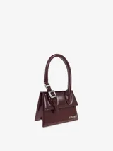 Bolso de mano estructurado de cuero color negro, con asa corta rígida, cierre con solapa y broche metálico dorado. Incluye correa fina desmontable y ajustable para usar como bandolera. Logotipo de la marca en letras doradas en la parte inferior frontal.