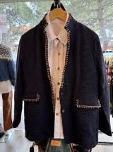 Blazer de textura tipo tweed en color azul marino, con bordados artesanales en hilo crudo a lo largo de los bordes frontales y en las solapas de los bolsillos. Presenta un diseño de corte recto y estructura flexible.