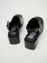 Zuecos de cuero vacuno con acabado en charol, color beige. Presentan un diseño clásico con taco cuadrado de madera de 5 cm y correa trasera ajustable con hebilla metálica.