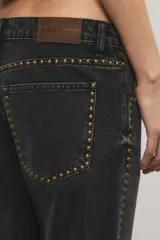 Jeans de denim negro de tiro medio y corte bootcut, con tachas metálicas doradas a lo largo de los laterales y en los bolsillos delanteros.