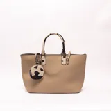 Cartera tipo shopper color beige con textura granulada, asas de mano y detalles con estampado animal print estilo vaca en las asas y en un pequeño monedero colgante.