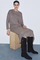 Bota de caña alta confeccionada en serraje, con diseño de estilo hípico. Presenta detalle de pespuntes verticales, tira decorativa con hebilla metálica en la parte superior de la caña y puntera redonda. Cuenta con suela plana de 2,5 cm y sistema de calce tipo pull-on.