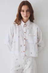 Camisa de corte recto y largo cropped, confeccionada en tela blanca con estampado de figuras deportivas pequeñas. Presenta cuello clásico, cierre frontal con botones de madera y costuras en hilo rojo a contraste. Incluye un bolsillo frontal en el pecho.