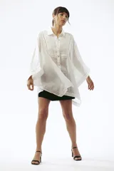 Camisa de lino blanca con corte oversize, cuello camisero y mangas amplias tipo kimono. Presenta detalles de costuras decorativas en el frente y cierre frontal con botones.