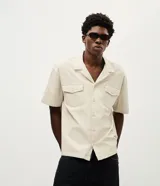 Camisa de corte boxy en color beige, con cuello inglés y mangas cortas. Presenta un diseño con lettering bordado en la espalda con la frase 'League of Almost Champions' y detalles gráficos en negro.