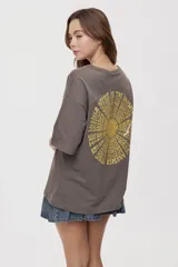 Remera de manga corta y cuello redondo, de corte oversized en color marrón, con texto bordado o estampado a lo largo del escote en el hombro izquierdo.