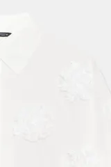 Camisa de tejido fluido color blanco, con cuello solapa, cierre frontal con botones y mangas amplias por debajo del codo. Presenta aplicaciones de flores en relieve a tono distribuidas por toda la prenda.