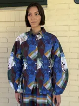 Camisa de popelina azul con estampado toile de jouy, cuello clásico y cierre frontal con botones. Presenta un diseño ajustado en la cintura mediante cordones laterales estilo corset y mangas largas abullonadas.