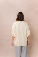 Buzo oversize color beige, con escote redondo, mangas cortas amplias de corte raglán y terminación frontal asimétrica en punta con aberturas laterales.