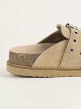 Zueco de gamuza color beige, con diseño de punta cerrada y talón descubierto. Presenta una tira ajustable sobre el empeine con hebilla metálica y ojales decorativos. Cuenta con una suela de corcho y base de goma antideslizante.