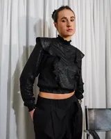 Camisa negra de lino con acabado brillante, cuello alto con volados, mangas largas abullonadas y detalles de volados frontales.
