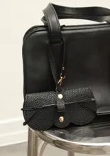 Estuche para lentes de cuero sintético negro con textura granulada y forma irregular. Tiene un cierre con broches dorados y una correa delgada con gancho para colgar de la cartera.