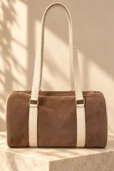 Cartera tipo baulito de cuero color marrón tostado, con doble asa larga de hombro, cierre superior y detalle de placa metálica con el logo de la marca en el centro.