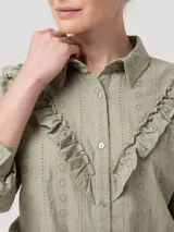 Blusa verde de algodón orgánico con cuello camisero, manga larga y cierre frontal de botones. Presenta volantes bordados que recorren el frente, la espalda y las sisas, además de detalles bordados en toda la prenda.