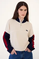 Sweater de punto acanalado con cuello alto y cierre frontal parcial. Presenta un diseño tricolor en bloques con cuerpo color crema, cuello y puños azul marino, y detalles en rojo en las mangas. Incluye una pequeña etiqueta con logo en el pecho.