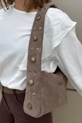 Cartera de hombro de gamuza color habano, con correa ancha decorada con tachas metálicas circulares de distintos tamaños. Incluye una placa metálica pequeña con el logo de la marca en el frente.