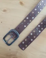 Cinto de cuero marrón con hebilla metálica de estilo envejecido y detalle de tachas metálicas en diferentes tonos en el extremo.