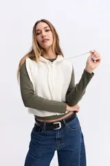 Chaleco de punto color crema con textura acanalada, diseño corto, cierre frontal y capucha con cordones ajustables.