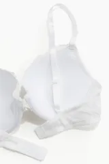 Soutien push-up blanco con aros y copas acolchadas, cubierto de encaje floral. Presenta breteles ajustables y un borde festoneado en la parte superior de las copas.