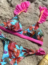 Conjunto de bikini con estampado floral en tonos naranja, marrón y verde sobre fondo celeste. El corpiño es triangular con volados en los breteles y la bombacha es regulable.