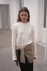 Sweater de punto color crema, con cuello redondo y cierre frontal parcial con botones. Presenta mangas largas con puños acanalados y detalle de volados en los extremos.