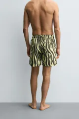 Bañador de hombre de largo medio, con estampado de cebra en tonos amarillo claro y negro/marrón oscuro. Presenta cintura elástica ajustable con cordón negro, bolsillos laterales y un bolsillo trasero de vivo.