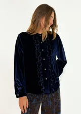 Camisa de terciopelo azul marino, con cuello redondo, cierre frontal de botones y detalle de volados verticales a lo largo de la botonera. Posee mangas largas con puños abotonados.