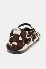 Zapato plano tipo bailarina con acabado de piel y estampado animal print de manchas marrones y blancas. Presenta una tira ajustable con hebilla metálica sobre el empeine y punta redonda.