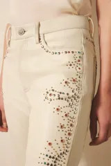 Pantalón de cuero ovino color crema, estilo jeanero con calce flare, adornado con bordados de tachas metálicas y piedras en tonos tierra y dorados a lo largo de las piernas.