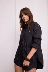 Camisa negra de algodón con bordados de pequeñas flores rosas. Presenta cuello clásico, cierre frontal con botones y mangas largas.