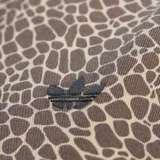 Campera deportiva Adidas modelo Firebird con cierre frontal, cuello alto y diseño estampado tipo animal print de jirafa en tonos marrones. Presenta las icónicas tres tiras de la marca en color negro a lo largo de las mangas y el logo del trébol bordado en el pecho.