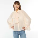 Camisa vaporosa color beige claro, con cuello alto y lazo al cuello, mangas largas abullonadas con puños elásticos.