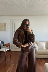 Campera bomber corta de cuero ovino marrón con detalles en napalan coñac y forro interior. Presenta cuello solapa, cierre frontal con cremallera de doble sentido, bolsillos laterales ribeteados, mangas largas con detalles de pasamanería cruzada y puños de piel sintética.