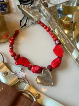 Collar artesanal con cadena de alpaca, compuesto por cuentas de coral rojo de forma irregular y cuentas redondas más pequeñas. Incluye un dije grande en forma de corazón de metal plateado.
