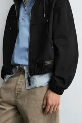 Cazadora negra de algodón con efecto lavado, corte relajado y capucha ajustable. Presenta cierre frontal de cremallera, bolsillos frontales tipo canguro y elásticos en puños y bajo.