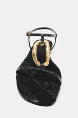Sandalia plana de color negro con textura efecto piel de serpiente. Presenta una tira central con un aplique metálico dorado curvo y cierre de pulsera con hebilla ajustable en el tobillo.