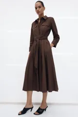 Vestido midi de estilo camisero color verde oscuro, con cuello solapa, escote en pico y cierre frontal con botones. Presenta manga larga, bolsillos laterales ocultos, solapas decorativas en el pecho y cinturón de lazada en el mismo tejido.
