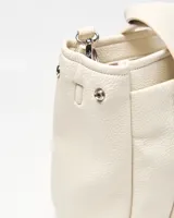Cartera mediana color crudo de simil cuero, con doble asa de mano, correa ajustable y desmontable, bolsillo frontal con botón a presión, detalles de herrajes plateados y dije colgante en forma de corazón. Cuenta con el logotipo de la marca grabado en la parte frontal.