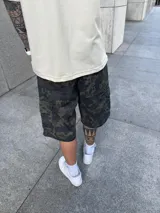 Bermuda tipo cargo con corte baggy, diseño de camuflaje en tonos verdes y negros, con bolsillos laterales y cierre frontal.
