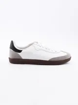 Zapatilla estilo urbano de color blanco, con refuerzo de gamuza gris en la puntera, suela de goma marrón y detalles en negro en el talón.