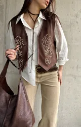 Chaleco de gamuza color marrón chocolate, con diseño sin mangas y escote en V. Cuenta con bordados en hilo claro y pequeños detalles de tachas en la parte frontal.