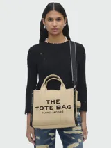 Bolso tote Marc Jacobs de lona en color beige, con el nombre del modelo estampado en contraste negro en el frente. Cuenta con doble asa superior, correa de hombro ajustable y extraíble, y cierres laterales.