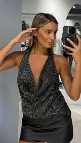 Top halter negro con acabado brillante y efecto drapeado en el escote. Presenta un diseño sin mangas con espalda descubierta y ajuste en el cuello.