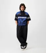 Remera de tricot liviano Blue Steel con corte boxy, cuello polo alto y cierre frontal parcial de un cuarto. Presenta diseño jacquard integral con gráficos y lettering en tonos azul, negro y blanco, mangas cortas y estilo de inspiración deportiva.