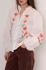 Camisa blanca de manga larga abullonada, con delicados bordados florales en tonos azules y verdes a lo largo de la botonadura frontal y en las mangas. Presenta cuello mao con reborde en contraste y cierre frontal con botones.