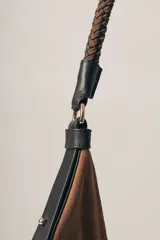 Cartera tipo hobo de gamuza color marrón, con correa trenzada de cuero negro y detalles laterales en cuero negro.