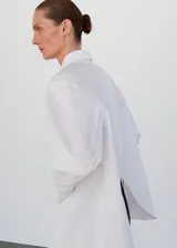 Camisa de algodón con diseño oversize y largo, cuello camisero clásico, cierre frontal con botones y mangas largas con puños abotonados. Presenta un diseño distintivo con espalda cruzada.