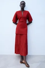 Vestido largo de punto color rojo, con cuello redondo, escote en V y manga corta. Presenta cierre frontal de botones a tono y una falda inferior de tejido combinado plisado.